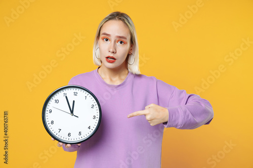 Young blonde caucasian smiling woman 20s with bob haircut bright makeup wearing casual basic purple shirt holding in hands pointing finger on clock isolated on yellow color background studio portrait.