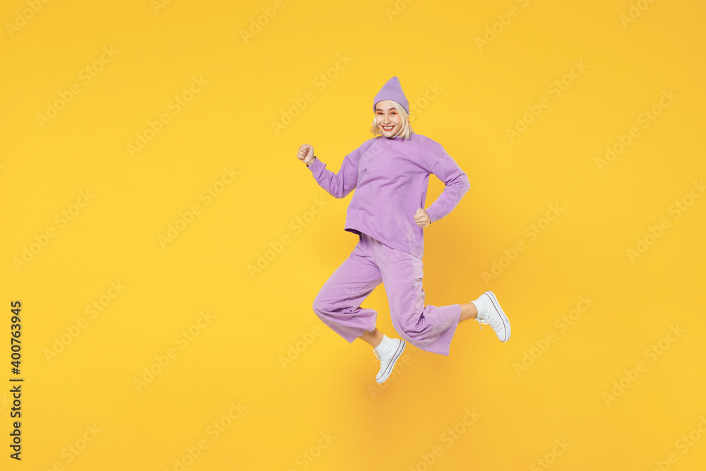 Full length of young caucasian excited fun woman 20s in casual basic purple suit beanie hat side view running jumping clenching fists look camera isolated on yellow color background studio portrait.
