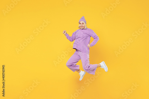 Full length of young caucasian excited fun woman 20s in casual basic purple suit beanie hat side view running jumping clenching fists look camera isolated on yellow color background studio portrait.
