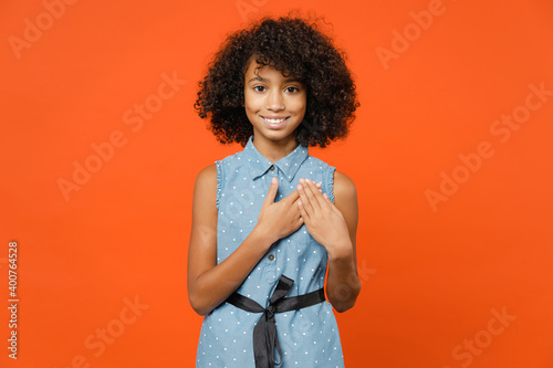 Photography Smiling pleasant little african american kid girl 12-13 years old in casual denim dress hold hands folded on heart isolated on orange background children studio portrait