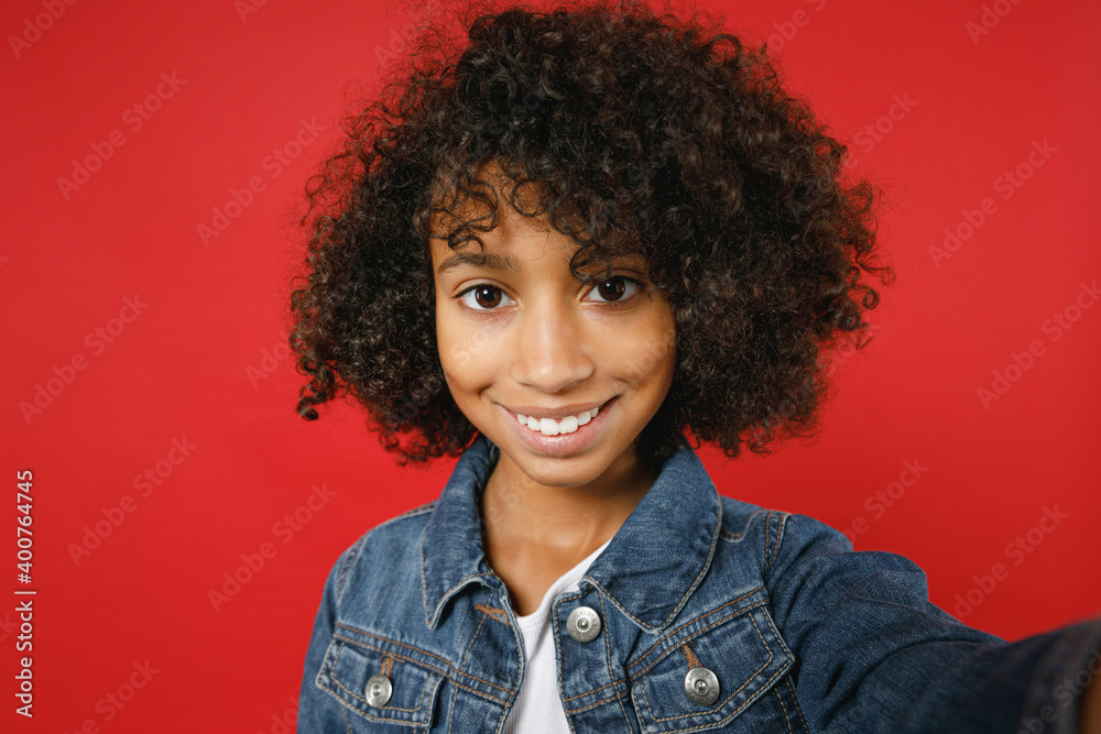Close up of smiling little african american girl 12-13 years old in denim jacket doing selfie shot on mobile phone isolated on red background children studio portrait. Childhood lifestyle concept. Close up of smiling little african american girl 12-13 years old in denim jacket doing selfie shot on mobile phone isolated on red background children studio portrait. Childhood lifestyle concept.