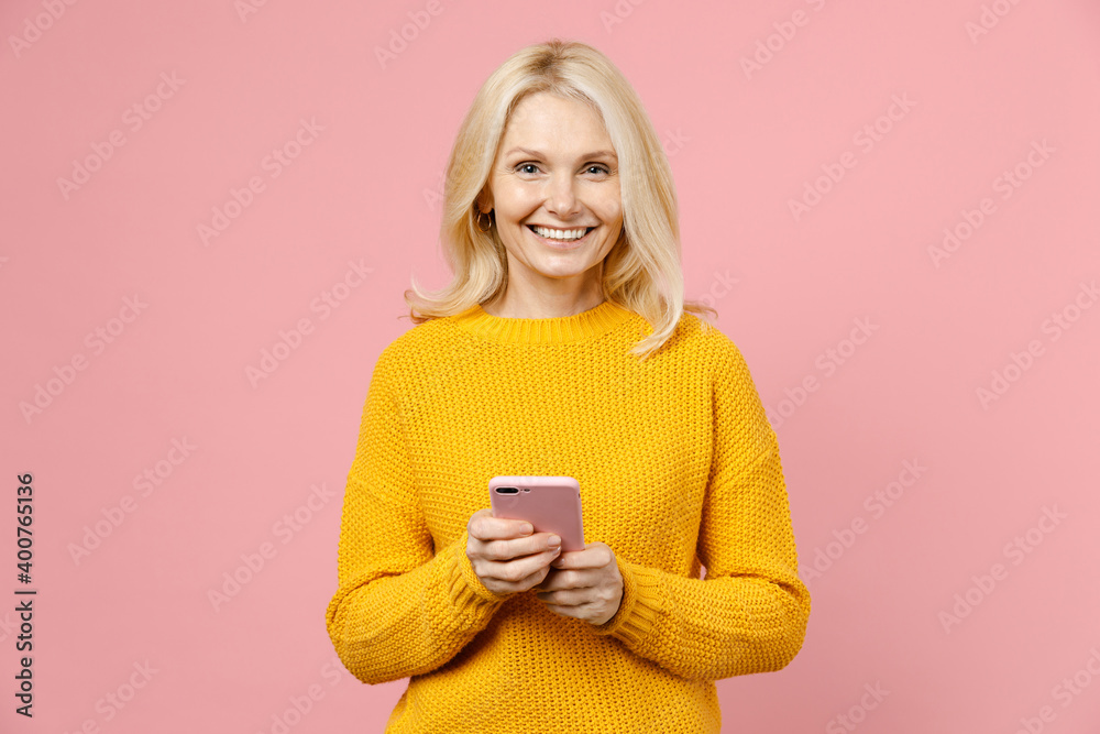Smiling elderly gray-haired blonde woman lady 40s 50s years old in yellow casual sweater standing using mobile cell phone typing sms message isolated on pastel pink color background studio portrait. Smiling elderly gray-haired blonde woman lady 40s 50s years old in yellow casual sweater standing using mobile cell phone typing sms message isolated on pastel pink color background studio portrait.