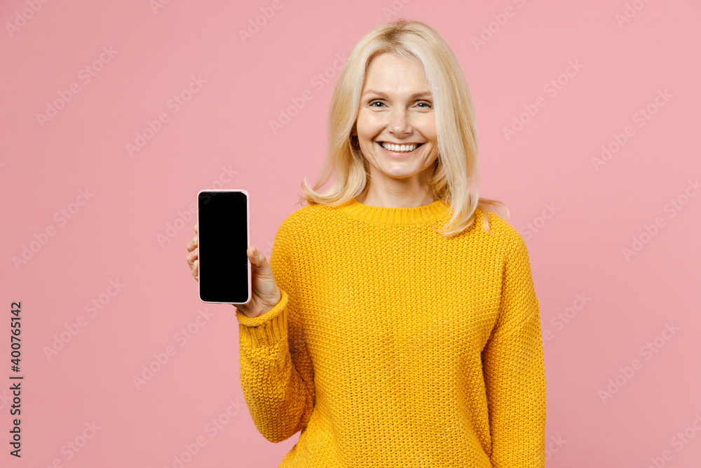 Smiling elderly gray-haired blonde woman lady 40s 50s years old in yellow casual sweater stand hold mobile cell phone with blank empty screen isolated on pastel pink color background studio portrait. Smiling elderly gray-haired blonde woman lady 40s 50s years old in yellow casual sweater stand hold mobile cell phone with blank empty screen isolated on pastel pink color background studio portrait.
