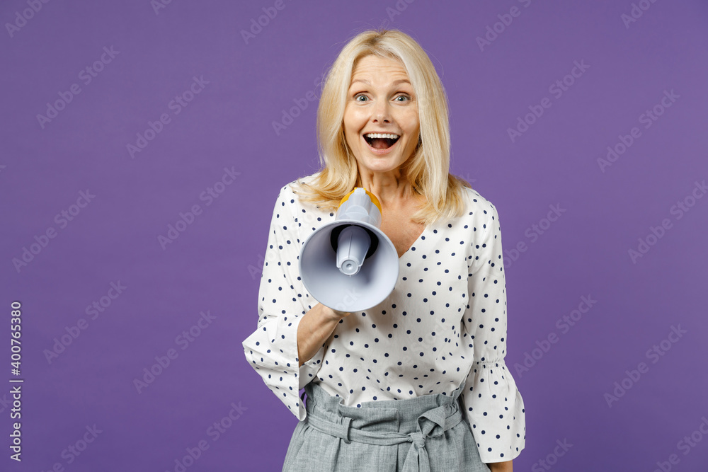 Excited funny elderly gray-haired blonde woman lady 40s 50s years old in white dotted blouse standing screaming in megaphone looking camera isolated on bright violet color background studio portrait. Excited funny elderly gray-haired blonde woman lady 40s 50s years old in white dotted blouse standing screaming in megaphone looking camera isolated on bright violet color background studio portrait.