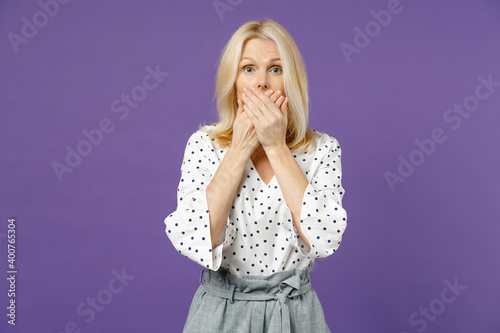 Shocked worried elderly gray-haired blonde woman lady 40s 50s years old wearing white dotted blouse covering mouth with hands looking camera isolated on bright violet color background studio portrait.