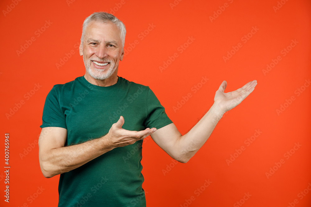 Smiling cheerful elderly gray-haired mustache bearded man wearing basic green t-shirt standing pointing hands aside on mock up copy space isolated on bright orange color background studio portrait. Smiling cheerful elderly gray-haired mustache bearded man wearing basic green t-shirt standing pointing hands aside on mock up copy space isolated on bright orange color background studio portrait.