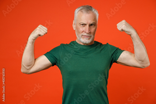 Foto Strong elderly gray-haired mustache bearded man wearing basic casual green t-shirt standing showing biceps muscles looking camera isolated on bright orange color wall background studio portrait