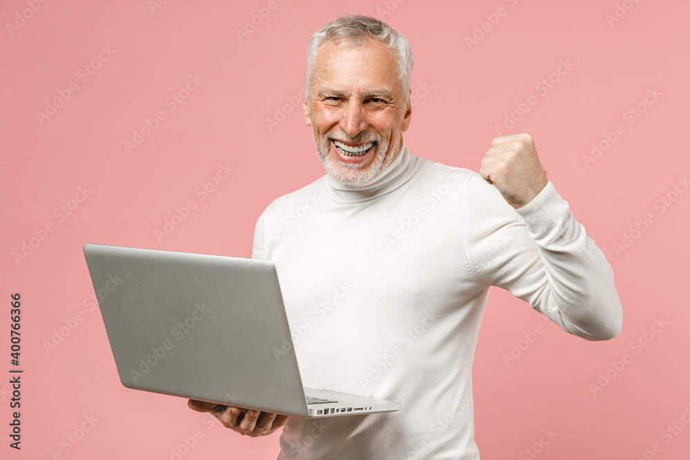 Happy elderly gray-haired mustache bearded man in basic casual white turtleneck standing working on laptop pc computer doing winner gesture isolated on pastel pink color background studio portrait. Happy elderly gray-haired mustache bearded man in basic casual white turtleneck standing working on laptop pc computer doing winner gesture isolated on pastel pink color background studio portrait.