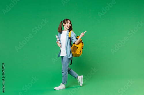 Full length side view of funny young woman student in blue shirt backpack hold notebooks pointing index finger aside isolated on green background. Education in high school university college concept.