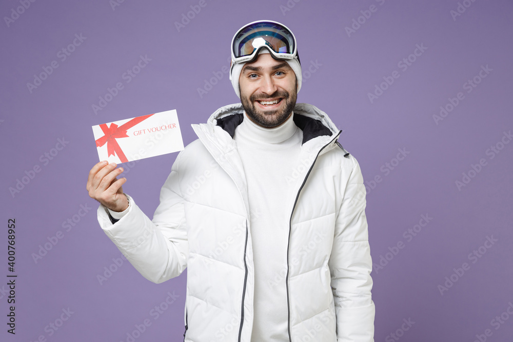 Funny skier man in warm white windbreaker jacket ski goggles mask hold gift certificate spend extreme weekend winter season in mountains isolated on purple background. People lifestyle hobby concept. Funny skier man in warm white windbreaker jacket ski goggles mask hold gift certificate spend extreme weekend winter season in mountains isolated on purple background. People lifestyle hobby concept.