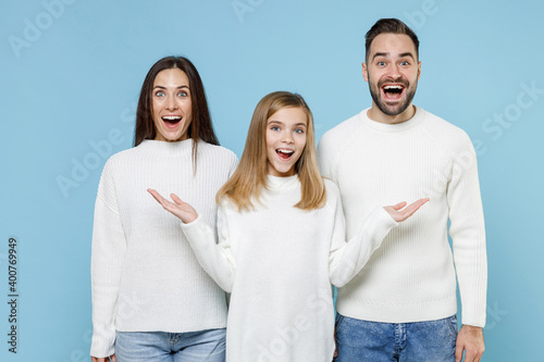 Excited young parents mom dad with child kid daughter teen girl in white sweaters keep mouth open spreading hands isolated on blue background studio portrait. Family day parenthood childhood concept.