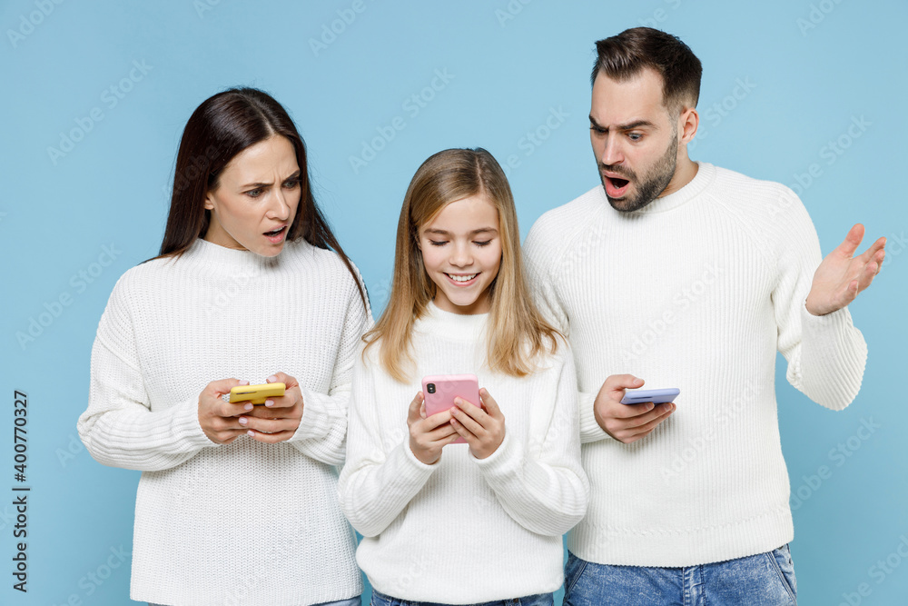 Shocked young parents mom dad with child kid daughter teen girl in white sweaters using mobile phone typing sms message isolated on blue background studio. Family day parenthood childhood concept. Shocked young parents mom dad with child kid daughter teen girl in white sweaters using mobile phone typing sms message isolated on blue background studio. Family day parenthood childhood concept.