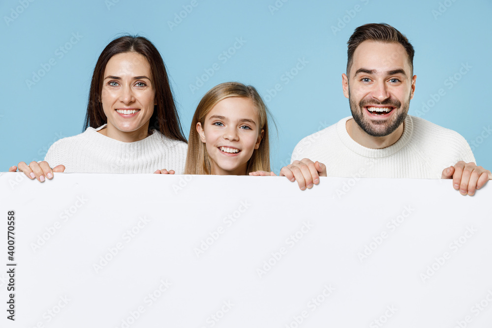 Funny young parents mom dad with child kid daughter teen girl in sweaters hold big white empty blank billboard mock up copy space isolated on blue background studio. Family day parenthood concept. Funny young parents mom dad with child kid daughter teen girl in sweaters hold big white empty blank billboard mock up copy space isolated on blue background studio. Family day parenthood concept.