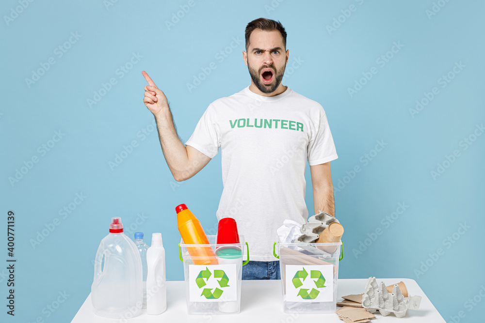 Worried young man in volunteer t-shirt stand near recycling stations sorting plastic paper trash point index finger up isolated on blue background. Voluntary free work assistance help grace concept. Worried young man in volunteer t-shirt stand near recycling stations sorting plastic paper trash point index finger up isolated on blue background. Voluntary free work assistance help grace concept.