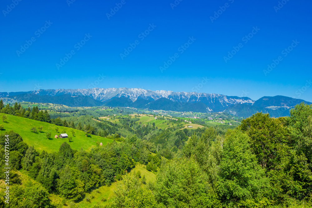 Stunning alpine landscape with green fields and high snowy Piatra Craiului mountains near Brasov. Mountain farm with old wooden house. Bran, Transylvania, Romania, Europe. 