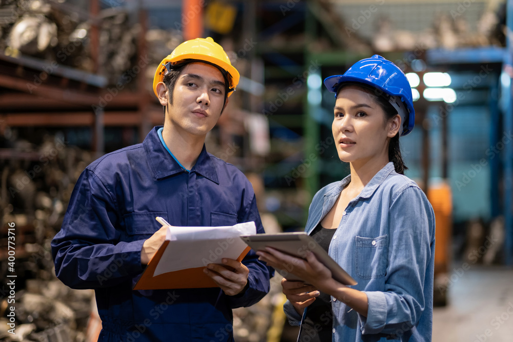 Fototapeta premium Engineer workers man wear a safety helmet writing on the clipboard in the automotive spare parts warehouse. Inspector the quality of the engine. Woman look at the tablet. Inspector, quality concept