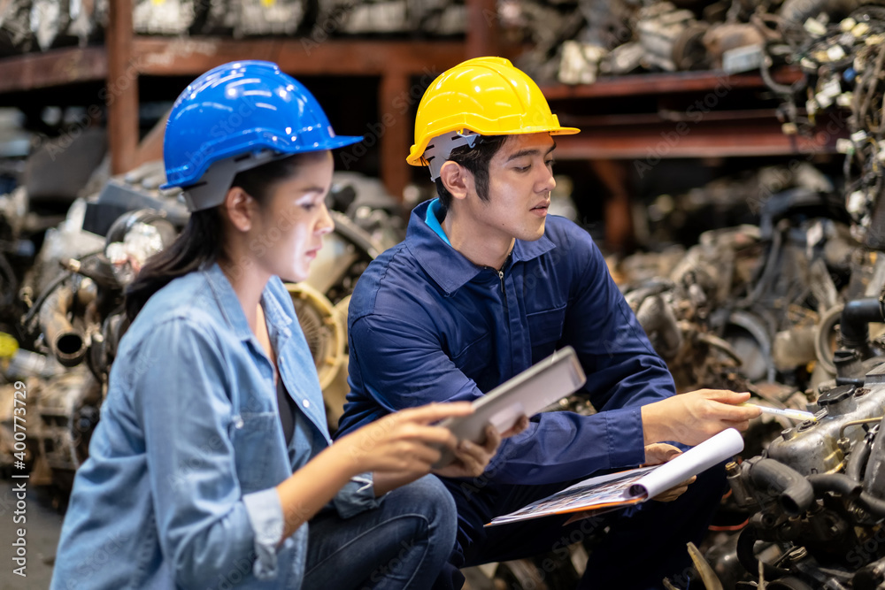 Engineer workers wear a safety helmet and sitting in the automotive spare parts warehouse. Inspector the quality of the engine. Woman look at the tablet. Maintainance and service concept. Engineer workers wear a safety helmet and sitting in the automotive spare parts warehouse. Inspector the quality of the engine. Woman look at the tablet. Maintainance and service concept.