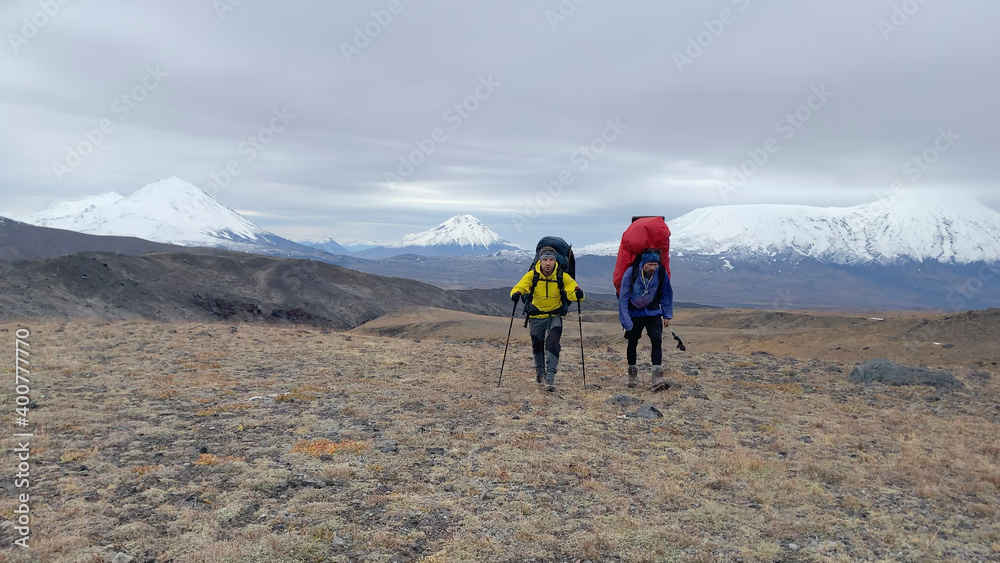 Two tourist hikers are walking along the tundra against the backdrop of the amazing volcanoes of Kamchatka. Two tourist hikers are walking along the tundra against the backdrop of the amazing volcanoes of Kamchatka.