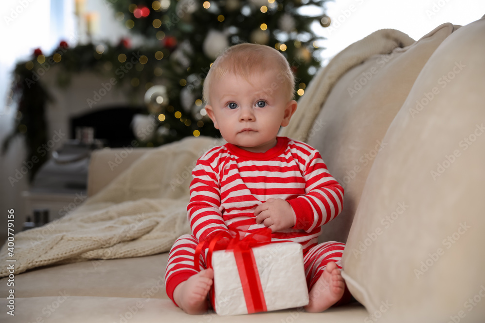 Cute baby in bright Christmas pajamas holding gift box on sofa at home Cute baby in bright Christmas pajamas holding gift box on sofa at home