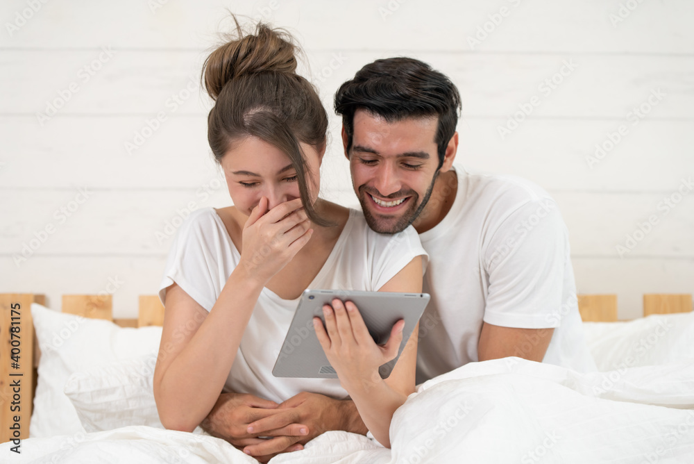 Happy young couple looking a tablet together and laughing while lying on the bed Happy young couple looking a tablet together and laughing while lying on the bed