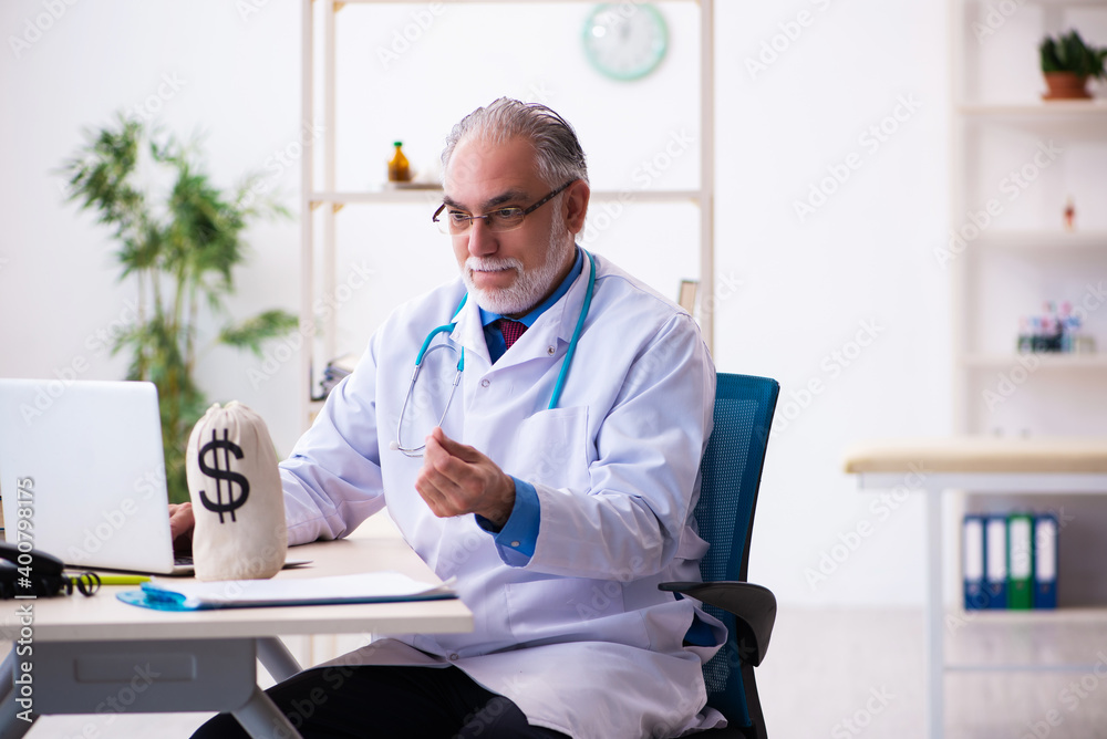 Obraz premium Old male doctor holding moneybag in the hospital