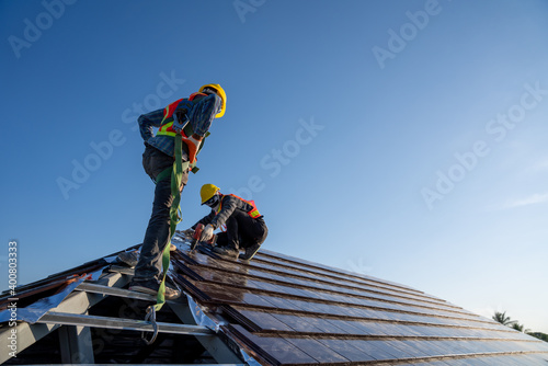 Construction worker wearing safety height equipment harness belt during working install new ceramic tile roof of building with Roofing tools electric drill used in the construction site.