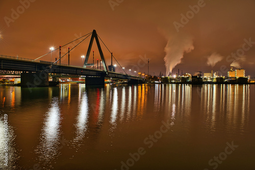 Hell erleuchtete Autobahnbrücke welche sich über den breiten Donau Fluss spannt mit qualmenden Industriegebäuden im Hintergrund photo