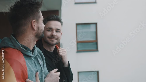 Proud lovely gay male couple standing on the balcony covered with pride rainbow flag. High quality 4k footage