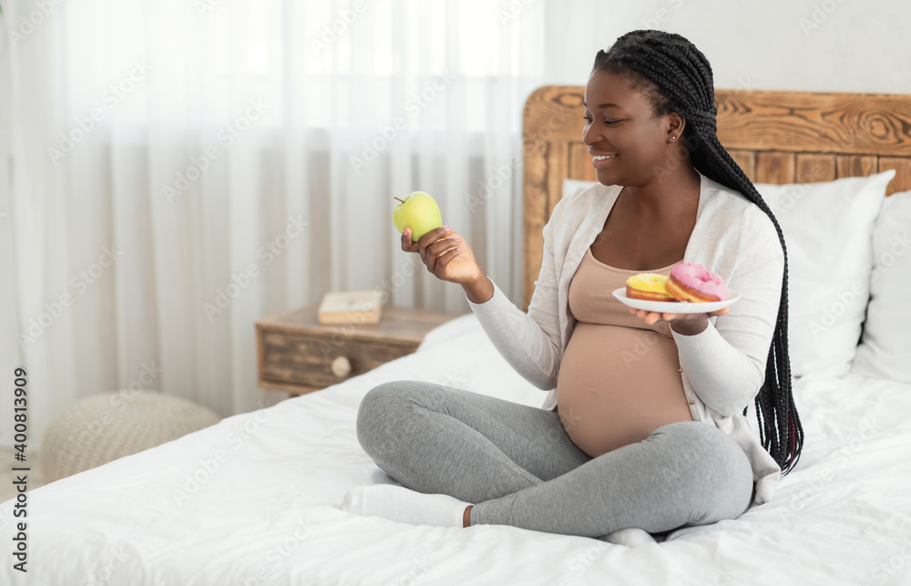 Prenatal Nutrition. Black Pregnant Woman Choosing Between Apple And Donuts At Home Prenatal Nutrition. Black Pregnant Woman Choosing Between Apple And Donuts At Home