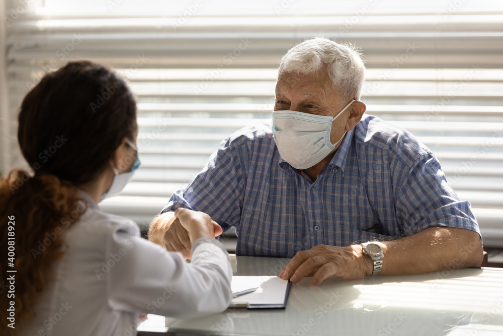 Obraz premium Smiling senior male patient in medical facial mask shake hand close health insurance deal with female doctor. Happy mature man client in facemask handshake woman GP at meeting in hospital.
