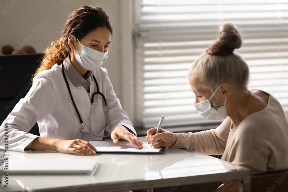 Mature female patient in medical facemask sign document close deal with GP or therapist in hospital. Elderly woman client put signature on contract make agreement about health insurance with doctor. Mature female patient in medical facemask sign document close deal with GP or therapist in hospital. Elderly woman client put signature on contract make agreement about health insurance with doctor.