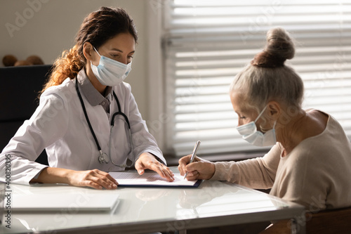 Mature female patient in medical facemask sign document close deal with GP or therapist in hospital. Elderly woman client put signature on contract make agreement about health insurance with doctor.