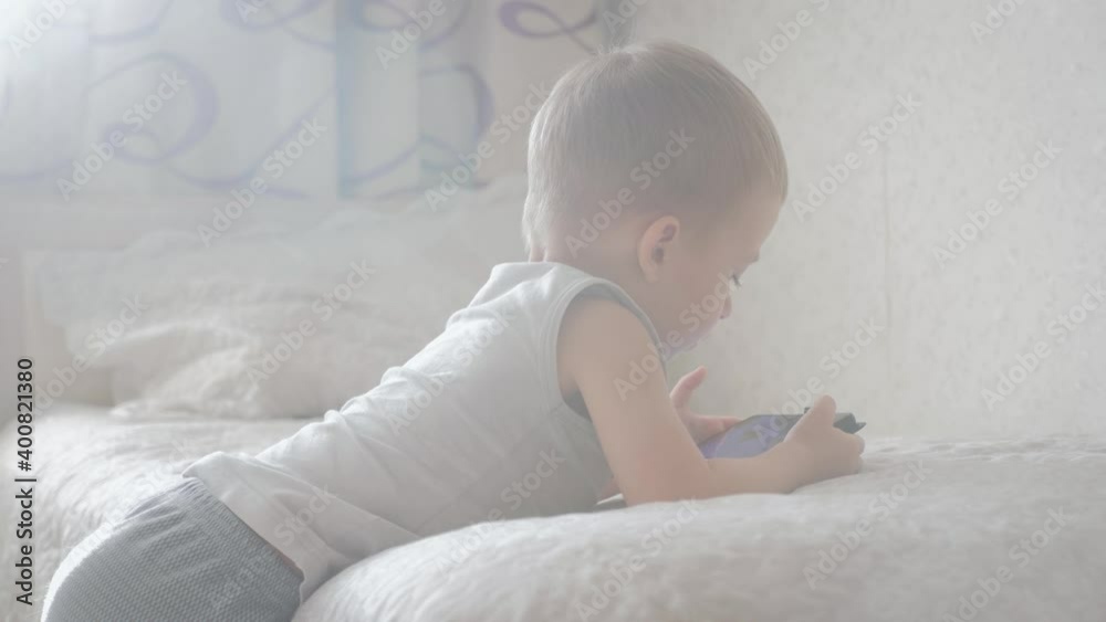 Little boy sitting on a bed and plays on the tablet in the room. Education and technology concept.
