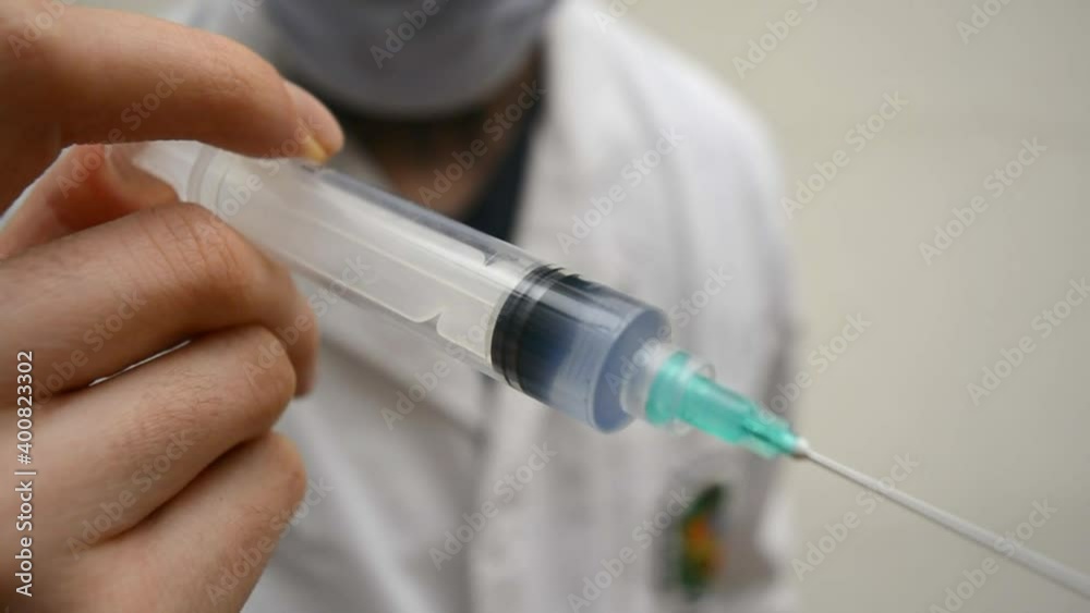Defocused doctor with white coat and mask, showing and injecting a vaccinDefocused doe with his hand in a disposable plastic syringe with blue liquid inside. Focused and detailed object in foreground. Defocused doctor with white coat and mask, showing and injecting a vaccinDefocused doe with his hand in a disposable plastic syringe with blue liquid inside. Focused and detailed object in foreground.