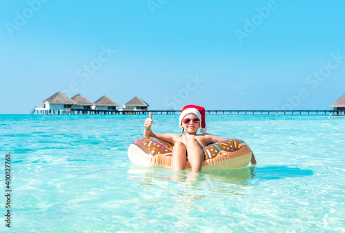 cute boy in a Santa hat on an inflatable donut in the ocean having fun and gives a thumbs up, the concept of Christmas and New year in exotic tropical countries and travel © klavdiyav