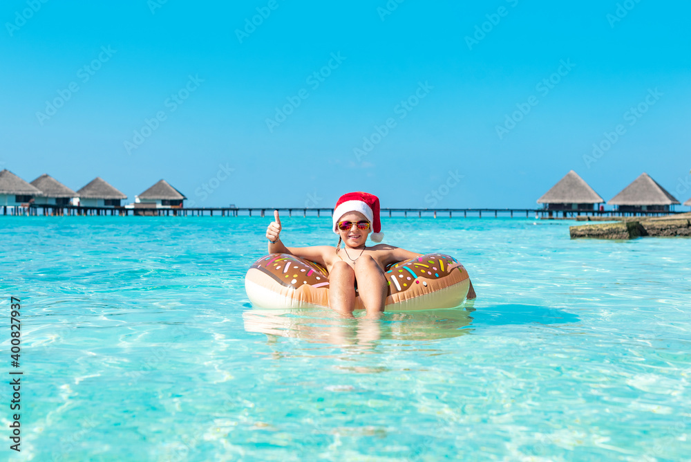 cute boy in a Santa hat on an inflatable donut in the ocean having fun and gives a thumbs up, the concept of Christmas and New year in exotic tropical countries and travel