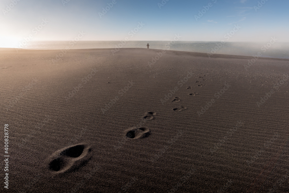 Lonely person silhouette standing far in background and footprints on the sand