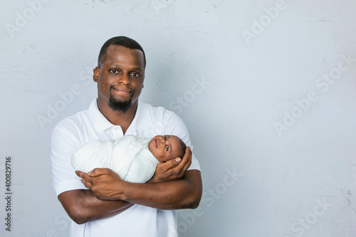 An attractive African-American new father is holding his newborn son and looking at the camera with a white-gray background.