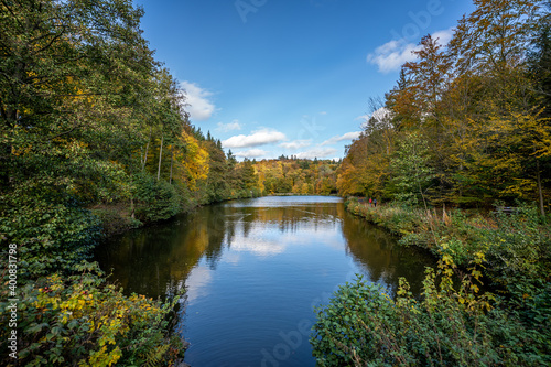 view of a lake in the autumn sunshine, lined with deciduous trees, which produce their leaves in beautiful yellow and red colors