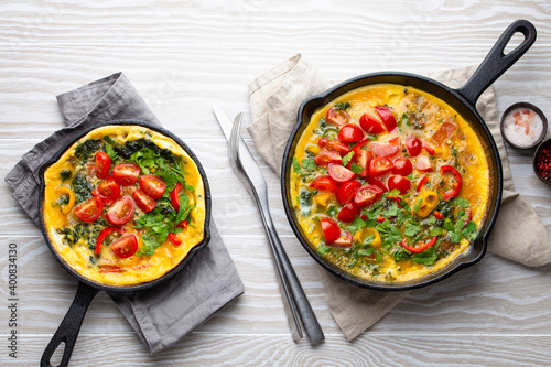Healthy frittata in cast iron pan with fried beaten eggs and seasonal vegetables on white rustic wooden background. Italian omelette with organic spinach, bell pepper, tomatoes, from above photo