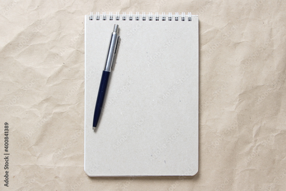 Gray notepad with white coiled spring and pen on a background of beige crumpled craft paper Gray notepad with white coiled spring and pen on a background of beige crumpled craft paper