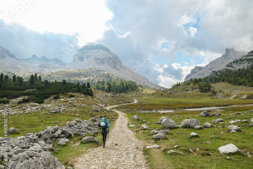 A woman with a hiking backpack hiking on a gravelled road in high Italian Dolomites. There is a dense forest at the foothill, and steep and sharp mountain chain in the back. Discovering and exploring photo