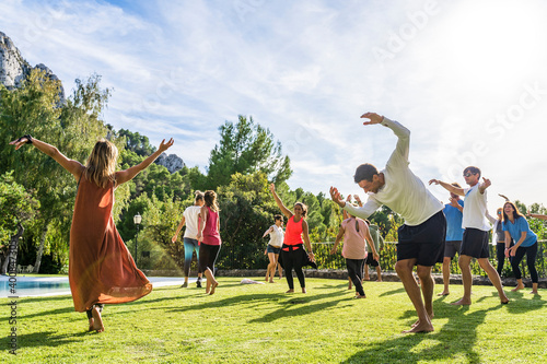 Mature female fitness instructor dancing with tourists at health retreat on grass against sky during sunny day