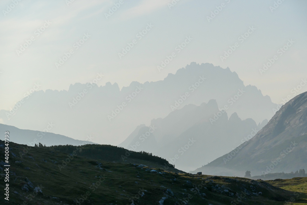 Obraz premium An early morning in Italian Dolomites. Lush green meadow in front. The valley is shrouded in morning haze. In the back there are high mountain chains. Sun slowly rising above the peaks. Golden hour