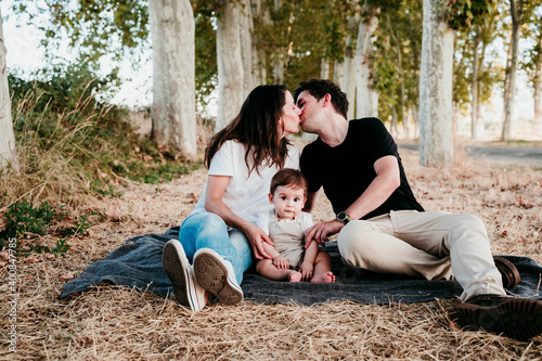 Parents kissing while sitting with baby bot on blanket outdoors photo