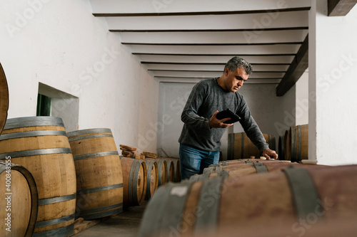 Winemaker holding digital tablet while examining wine barrels at cellar photo