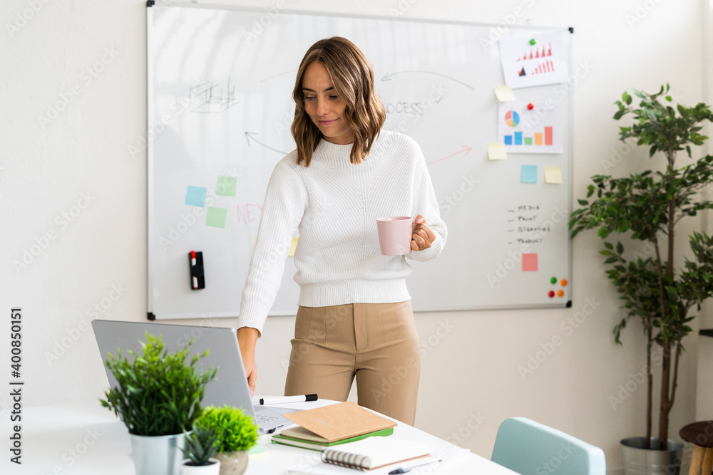 Young businesswoman with coffee cup using laptop while standing at office Young businesswoman with coffee cup using laptop while standing at office