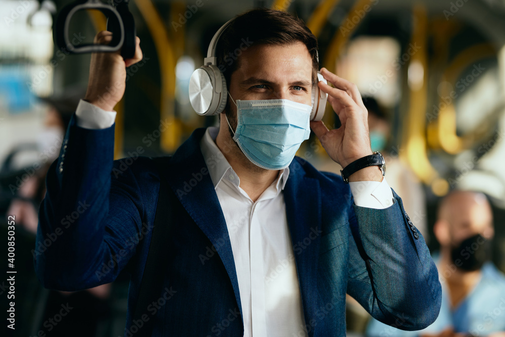 Businessman with face mask listening music over headphones while traveling to work by bus.