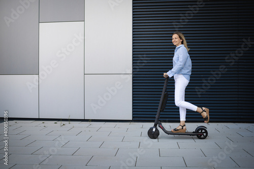 Mid adult woman driving electric push scooter on footpath by metal wall photo