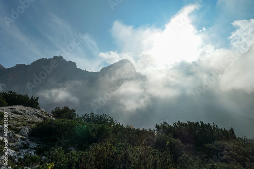 A view on a high and sharp mountains towering above the ground level. The peaks are shrouded with a few clouds. Sunbeams coming through the clouds. Stony  steep mountain walls. Discovering
